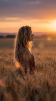 Young woman with long wavy hair standing in golden wheat field during sunset with warm bokeh lights and dramatic sky for lifestyle photography.