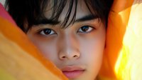 Young Asian man with dark hair and brown eyes surrounded by vibrant orange and yellow fabric creating warm lighting effects for fashion and beauty concepts.