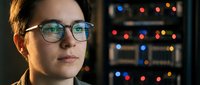 Young Asian female IT specialist wearing glasses working late in server room with colorful network equipment lights reflecting on lenses.