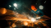 Welder in protective mask working with metal, creating bright orange sparks and smoke in dark workshop environment.