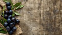 Fresh ripe black olives with green leaves on rustic burlap sack against dark wooden background, copy space for Mediterranean food styling.