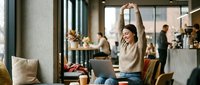 Young woman stretching arms above head while sitting at cafe table, taking break from laptop work in modern coffee shop with warm natural lighting.