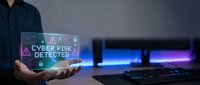 Male professional in dark business attire holding holographic display showing cyber risk alert with warning symbol, modern computer workstation with neon lighting in blurred background, cybersecurity threat notification concept.