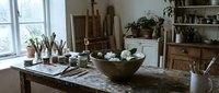 Rustic artist studio interior with wooden table, paint brushes, ceramic bowl with white flowers, easels, and shelves with plants in natural light.