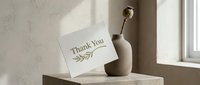 Elegant thank you card with botanical leaf design leaning against a minimalist beige ceramic vase with dried flower on a stone surface.