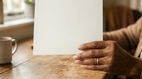 Senior man with dark skin tone holding blank white card at wooden table with coffee cup, suitable for business messaging, greeting cards, and promotional campaigns.