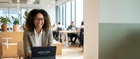 Happy mixed-race businesswoman with curly hair working on laptop in modern office space, smiling at camera while colleagues collaborate in background.