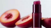 Fresh cut ripe nectarine halves next to burgundy lip balm on white background, natural cosmetics concept showing fruit-inspired beauty products.