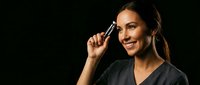 Young Hispanic woman applying mascara with makeup brush smiling at camera against black background for beauty and cosmetics advertising campaigns.