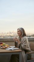Mature woman enjoying morning coffee on rooftop terrace with city skyline view, relaxing breakfast moment during golden hour sunrise.