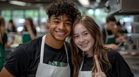 Young diverse restaurant workers in black uniforms and aprons smile warmly at camera. Friendly coworkers showcase authentic workplace camaraderie in casual dining setting.