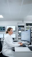 Female scientist in white lab coat analyzing data on computer monitors in modern laboratory control room for research and technology applications.