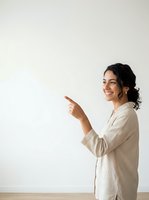 Young woman with dark curly hair and olive complexion wearing a beige linen shirt, smiling while pointing to the left against a neutral white wall background. Suitable for business, advertising, and promotional materials.