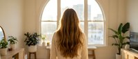 Young woman with long brown hair standing by large arched window in bright modern living room with plants and natural light streaming in.