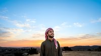 Middle Eastern man wearing traditional pink and white headwrap and olive jacket standing in desert landscape at sunset, gazing toward horizon with mountains in background.