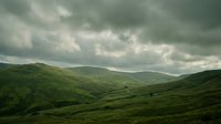 Rolling green hills under dramatic cloudy sky, rural valley landscape with mountains in distance, moody weather conditions.