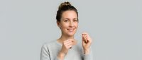 Woman with brown hair in bun, Caucasian ethnicity, wearing gray sweater, smiling with hands near face in cheerful pose against neutral background.