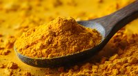 Ground turmeric powder on wooden spoon with scattered yellow spice on surface, macro food photography showing vibrant golden color and texture of natural seasoning.