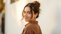 Young woman with messy bun hairstyle wearing brown jacket looking over shoulder with warm smile in soft natural lighting for lifestyle photography.