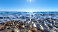 Sparkling ocean waves ripple under bright sunlight with sun star effect, close-up view of clear blue seawater surface showing detailed water texture and reflections.