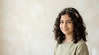 Young woman with long curly dark hair and South Asian ethnicity wearing a delicate pearl headpiece and sage green top, smiling at camera against neutral beige background for portrait and lifestyle use.