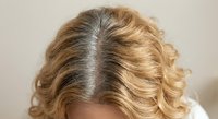 Blonde woman showing gray hair roots growing out from center part revealing natural hair color transition and regrowth pattern on wavy styled hair.