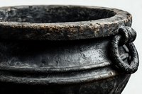 Ancient cast iron cauldron with rustic metal handle and weathered rim showing patina and texture against white background.