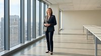 Mature blonde woman in professional black blazer and trousers standing in modern high-rise office corridor with floor-to-ceiling windows overlooking cityscape, suitable for corporate business and leadership concepts.