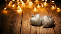 Two silver glitter heart ornaments on rustic wooden surface with warm bokeh fairy lights in background, romantic holiday decor.