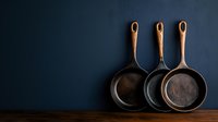 Cast iron skillets arranged in row on wooden surface against dark blue wall, rustic kitchen decor with vintage cookware showing minimalist composition.