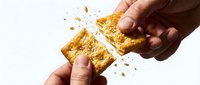Hands breaking golden crispy snack cake with crumbs scattering against white background, food sharing moment.