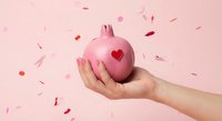 Hand holding pink pomegranate with red heart symbol surrounded by falling petals on pink background for Valentine's Day and love concepts.