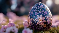 Ornate Easter egg with delicate floral pattern in blue and pink, decorated with intricate swirls and flowers, placed on grass with soft pink blossoms in background.