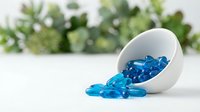 Blue capsules spilling from white ceramic bowl on clean surface with green plants in background for pharmaceutical and healthcare concepts.