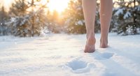 Barefoot woman walking through fresh snow leaving footprints in winter landscape with warm golden sunlight filtering through snowy trees creating magical atmosphere.