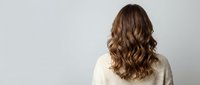 Young woman with long wavy brown hair from behind wearing cream sweater against light gray background for hair care and beauty concepts.