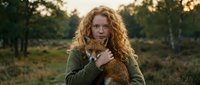 Young Caucasian woman with curly red hair holding small brown dog outdoors during golden hour sunset in natural park setting with warm lighting.