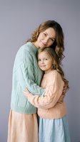 Loving mother and daughter embracing in cozy knit sweaters against neutral background. Warm family portrait showcasing maternal bond and affection between generations.