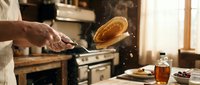 Chef flipping pancake in rustic kitchen with golden morning light streaming through window creating warm cooking atmosphere for breakfast preparation.