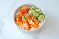 Fresh salmon sashimi and cucumber slices on white rice in ceramic bowl, garnished with sesame seeds, served on marble surface. Asian cuisine.