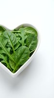 Fresh spinach leaves in heart-shaped white bowl on white background. Healthy eating, nutrition, and diet concept for food and wellness projects.