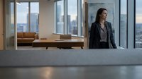 Woman in business attire standing in modern high-rise office with floor-to-ceiling windows overlooking city skyline. Corporate professional environment.