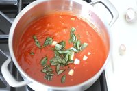 Fresh homemade tomato soup garnished with basil leaves and cream in white pot on stovetop, overhead view showing rich red color and smooth texture.