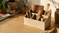 Wooden desk organizer with bamboo utensils and craft supplies on natural wood surface with plants in warm sunlight for home office workspace organization.