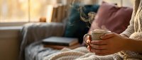 Woman in cozy knit sweater holding warm coffee mug in comfortable living room with soft pillows and blankets creating hygge atmosphere for relaxation.