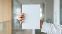 Young adult male with medium skin tone holding blank white paper sheet in modern office environment for mockup and branding purposes.