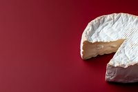 Soft ripened white mold cheese wheel with missing slice on deep red background, showing creamy texture and characteristic rind. Food photography concept.