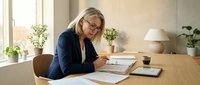 Middle-aged woman with blonde hair and glasses, wearing a dark blazer, working at a wooden desk with documents and tablet, surrounded by potted plants and natural light from window.