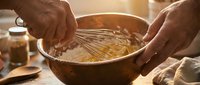 Hands whisking ingredients in wooden bowl for homemade baking preparation in warm kitchen setting with natural lighting and cooking utensils nearby.