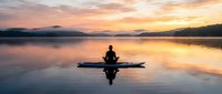 Silhouette of kayaker paddling on calm lake at golden sunrise with mountain reflections in serene water creating peaceful outdoor recreation scene.
